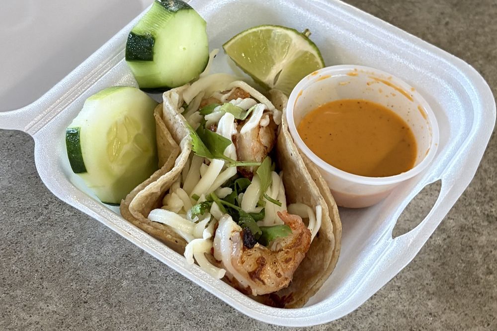 A shrimp street taco with cheese and cilantro, served with cucumber slices, a lime wedge and hot sauce